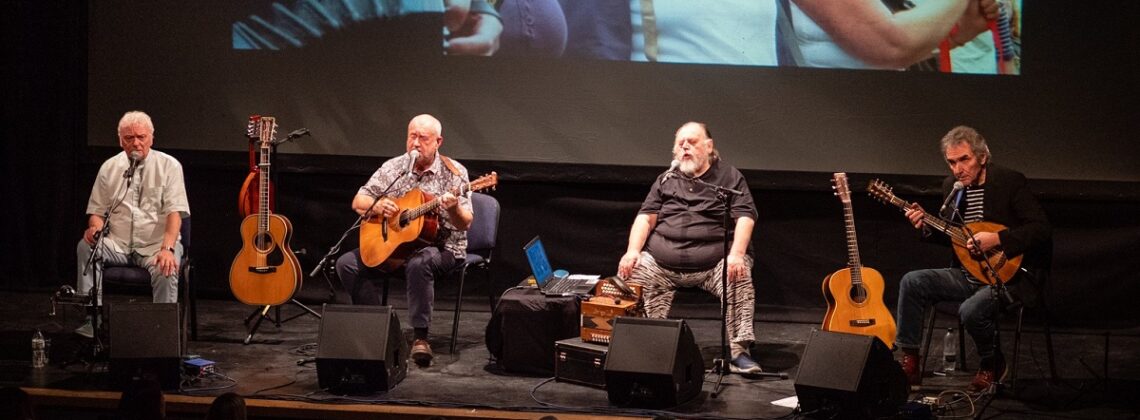 The Pitmen Poets perform on stage with footage from a protest playing behind them.