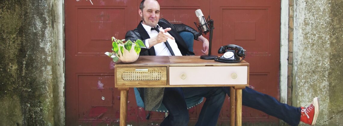 Comedian Phil Ellis sits at a desk in front of a red garage door with a plant, microphone and a telephone. He is wearing a suit and has three legs.