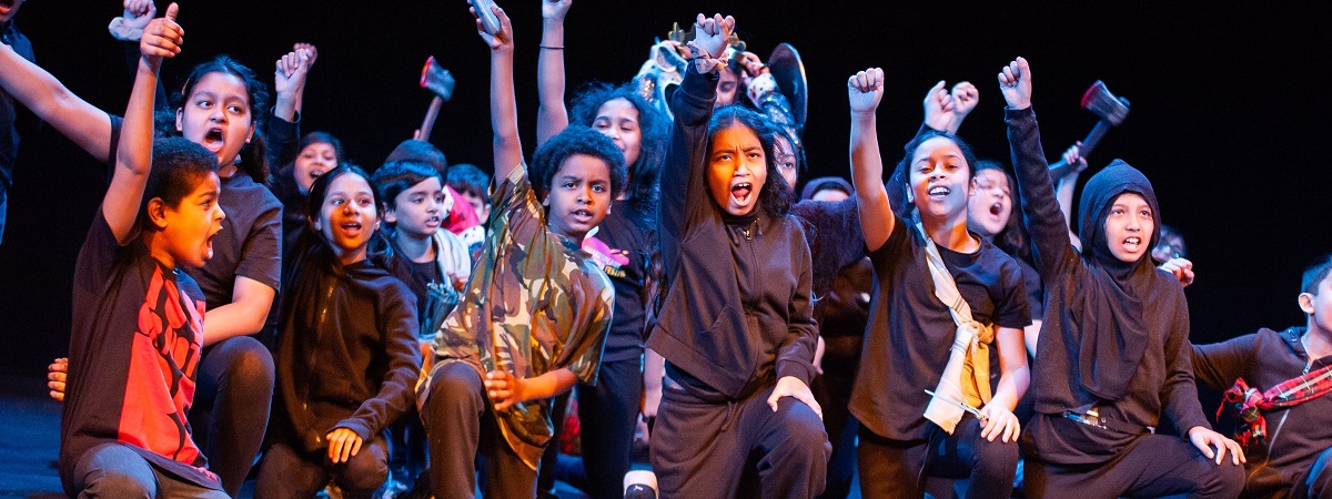 A group of school children on one knee with an arm in the air in a promotional image for the Coram Shakespeare Schools Festival.