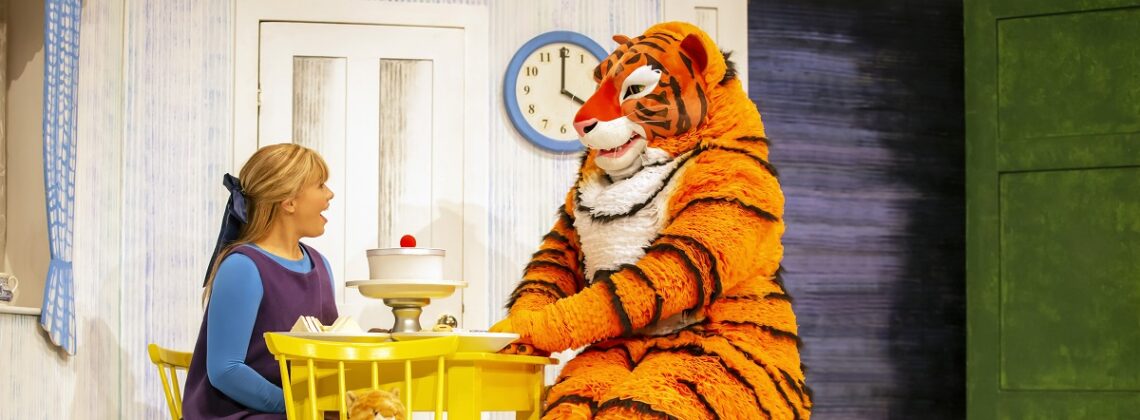 A girl sits at a table with someone dressed as a tiger in a production shot from The Tiger Who Came To Tea