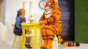 A girl sits at a table with someone dressed as a tiger in a production shot from The Tiger Who Came To Tea