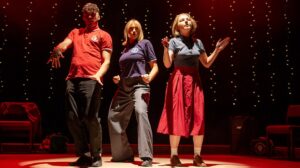 Three people wearing tops with a 'Northern Soul - Keep The Faith' logo dance in a production shot from John Godber's Do I Love You?