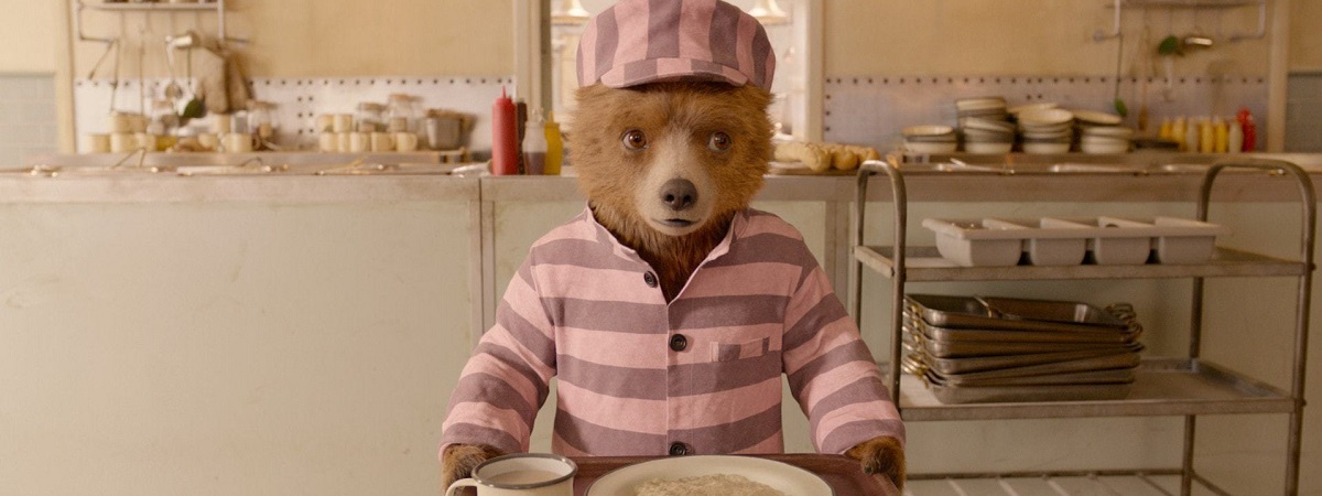 Paddington wearing a jail outfit whilst carrying a drink and a bowl on a tray in a still from Paddington 2.