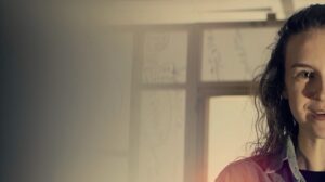 A women looks straight ahead with only the right side of their face visible with a door and wall in the background in a promotional image for Tiny Fragments of Beautiful Light.