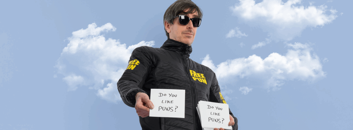 Comedian Darren Walsh stands wearing a jacket with 'Free Pun' on it holding two pieces of paper reading 'Do You Like Puns?'