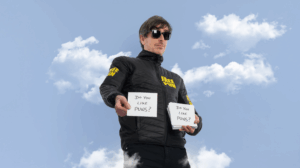 Comedian Darren Walsh stands wearing a jacket with 'Free Pun' on it holding two pieces of paper reading 'Do You Like Puns?'