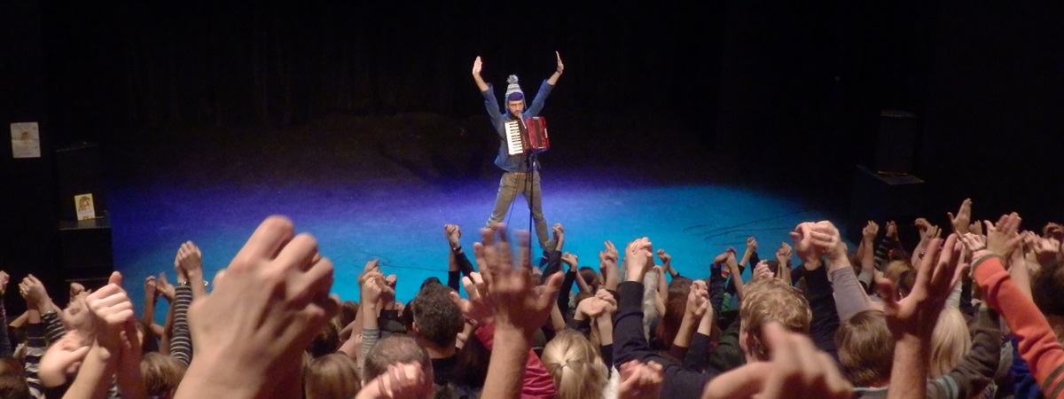 A comedian stands on stage as audience members have hands in the air in an image for Comedy Club 4 Kids.