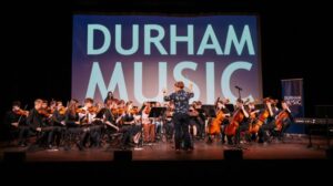 A photograph of a group of young people playing music in front of a screen which reads 'Durham Music'.